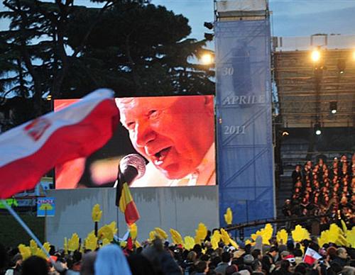 Peregrinos se reúnem em frente a uma tela que mostra imagens do Papa João Paulo II, em Roma